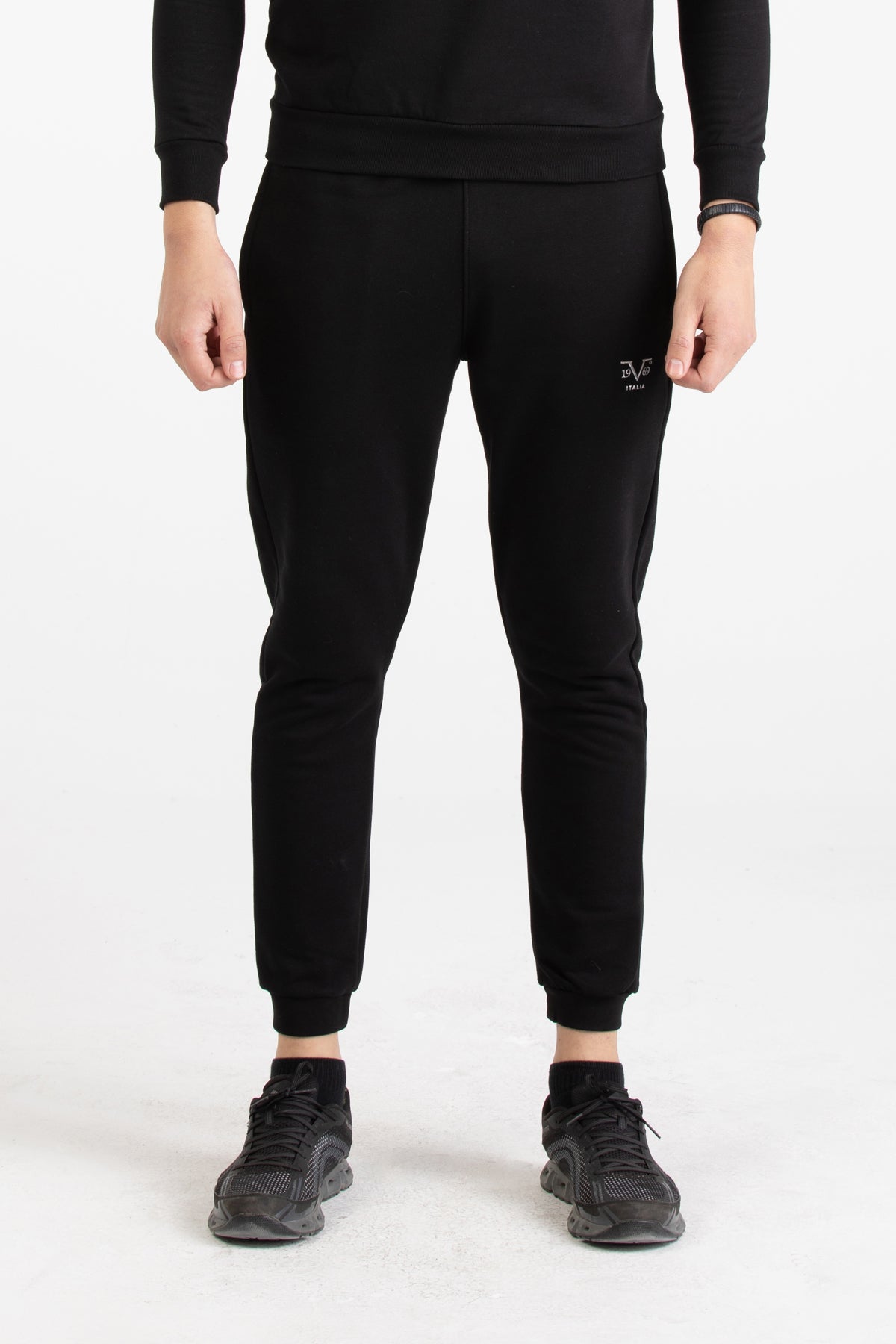 Full-body view of a man wearing a black Versace 19.69 tracksuit set with silver 19V69 linear branding on the joggers.