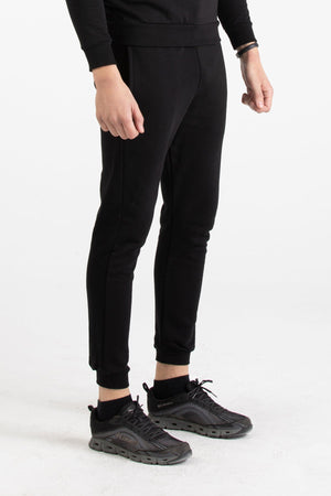 Full-body view of a man wearing a black Versace 19.69 tracksuit set with silver 19V69 linear branding on the joggers.