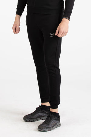 Full-body view of a man wearing a black Versace 19.69 tracksuit set with silver 19V69 linear branding on the joggers.